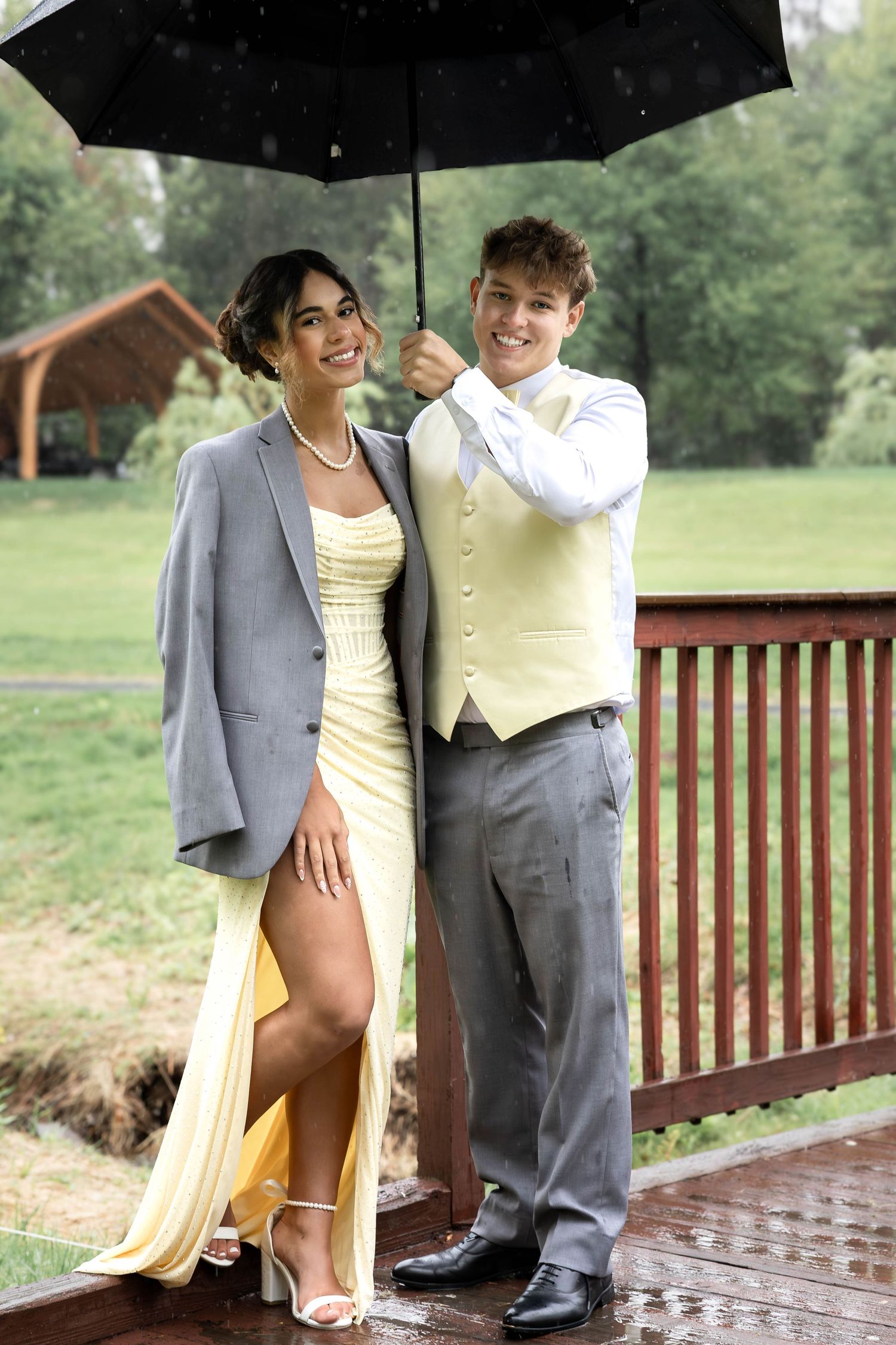 Young couple in formal wear sharing umbrella in rain, girl in yellow dress with gray blazer, boy in vest and slacks.