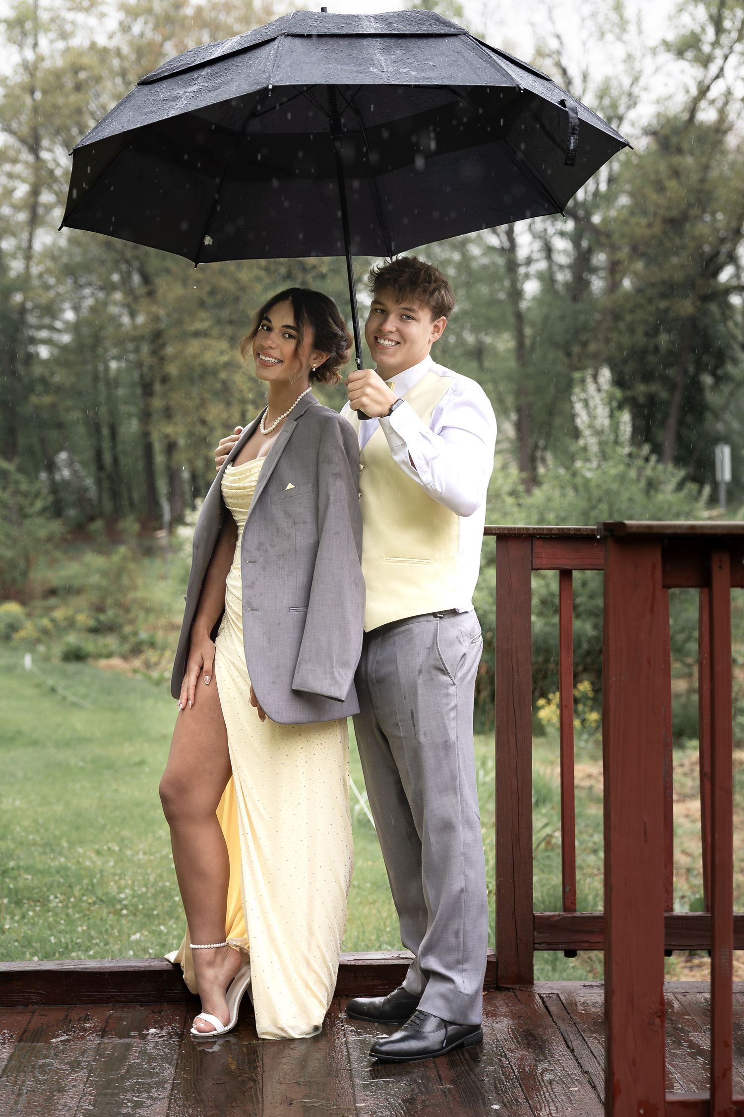 Couple in formal prom attire posing on rainy deck, woman in yellow gown, man in gray suit holding black umbrella.