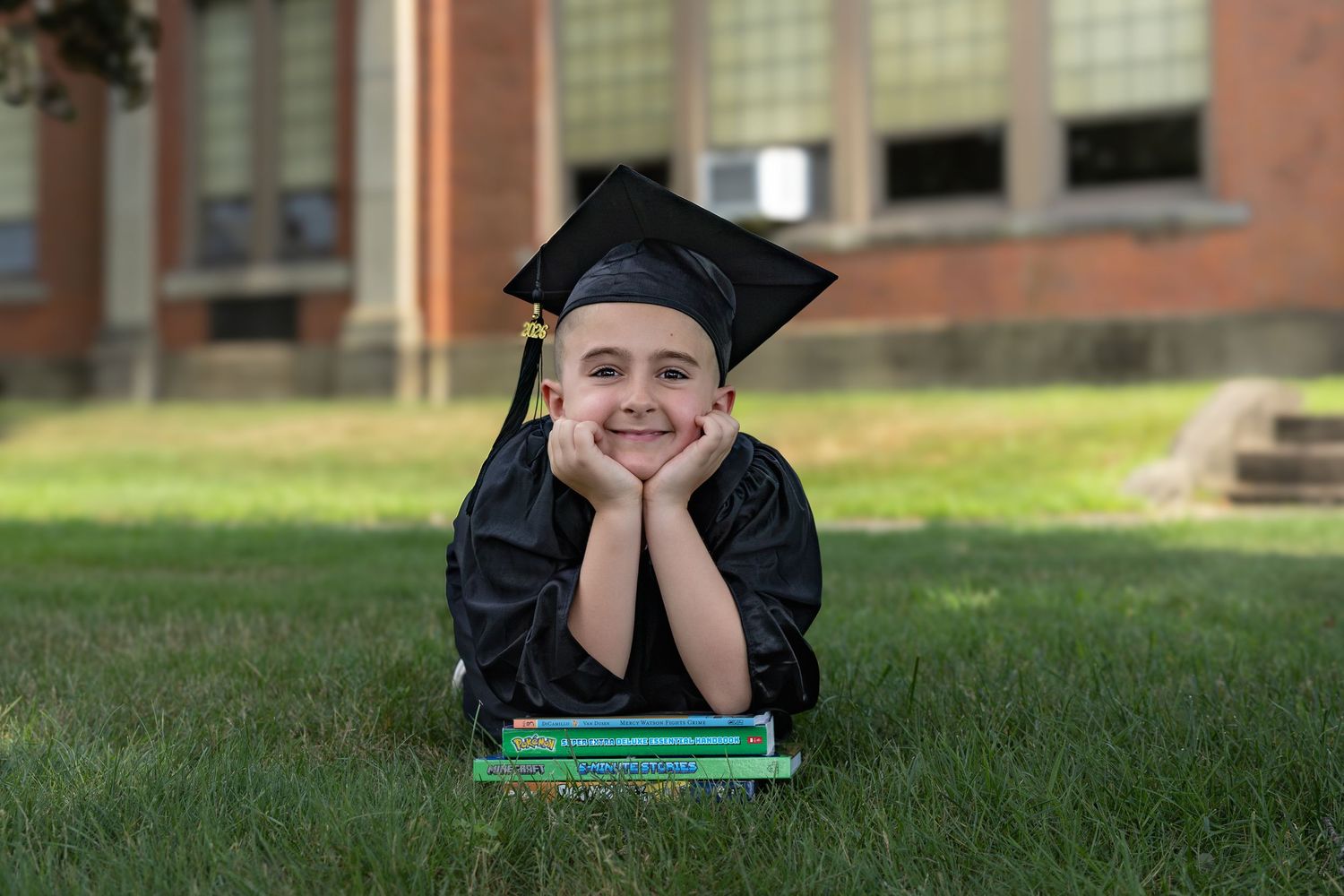 Young child in graduation cap and gown lying on grass, smiling with books, in front of a brick building.