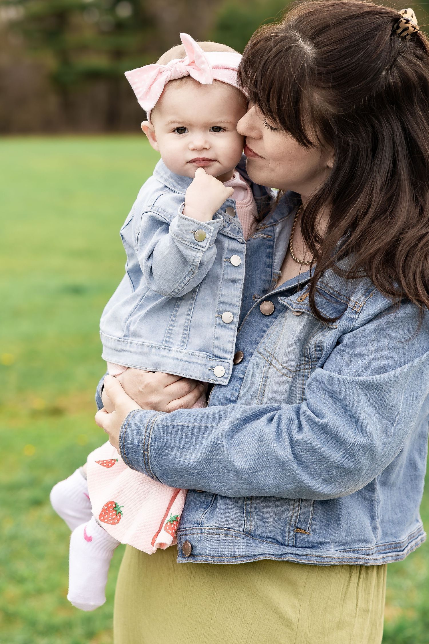 A mother in a denim jacket kisses her baby girl wearing a pink bow headband outdoors on green grass.