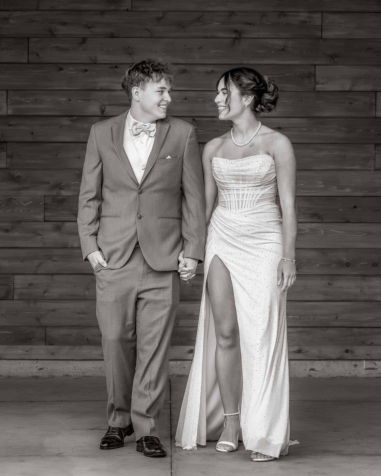 Black and white photo of a couple in formal attire holding hands and smiling at each other against a wood wall.