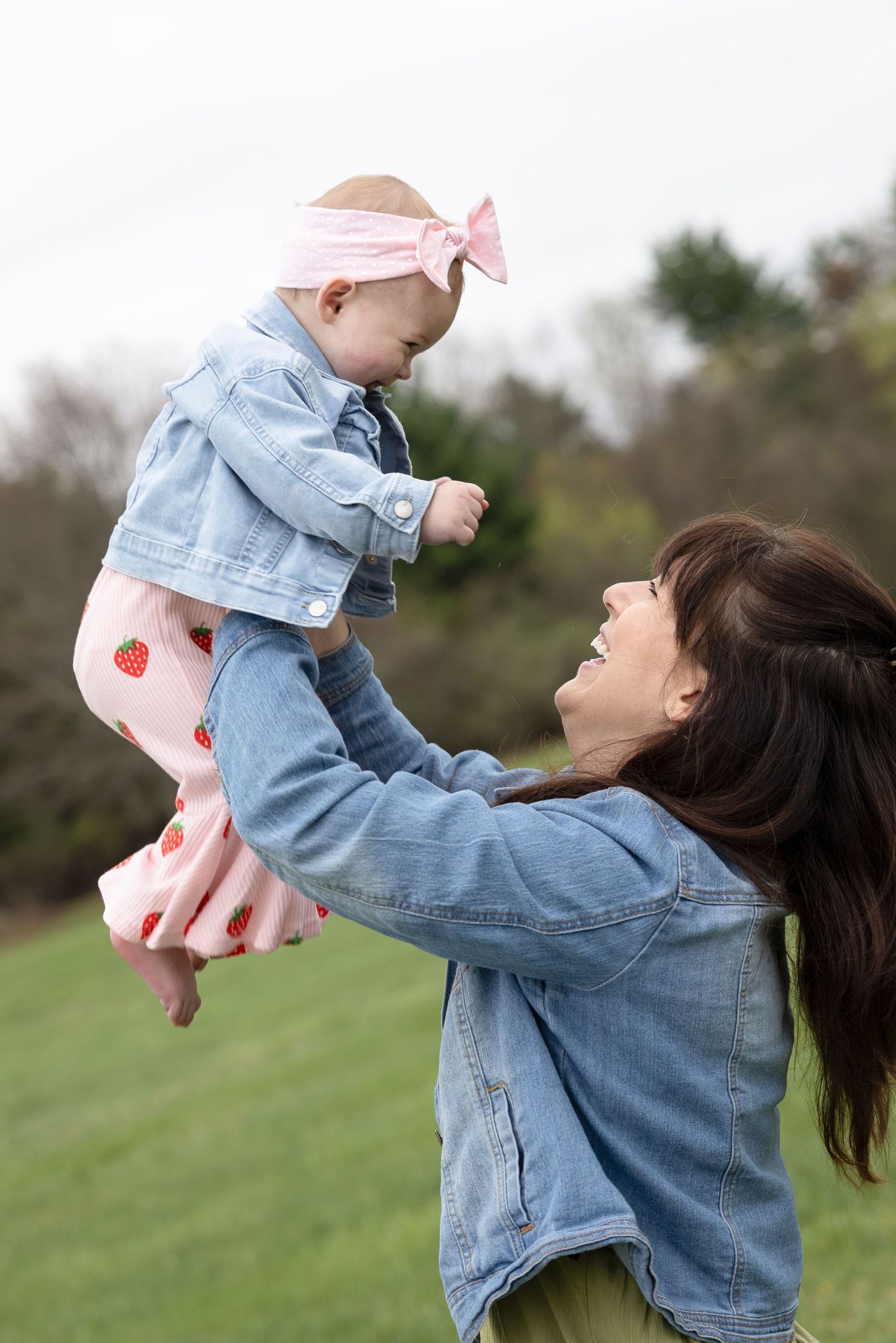 A smiling mother lifts her baby girl wearing a pink bow and strawberry outfit in a green outdoor setting.