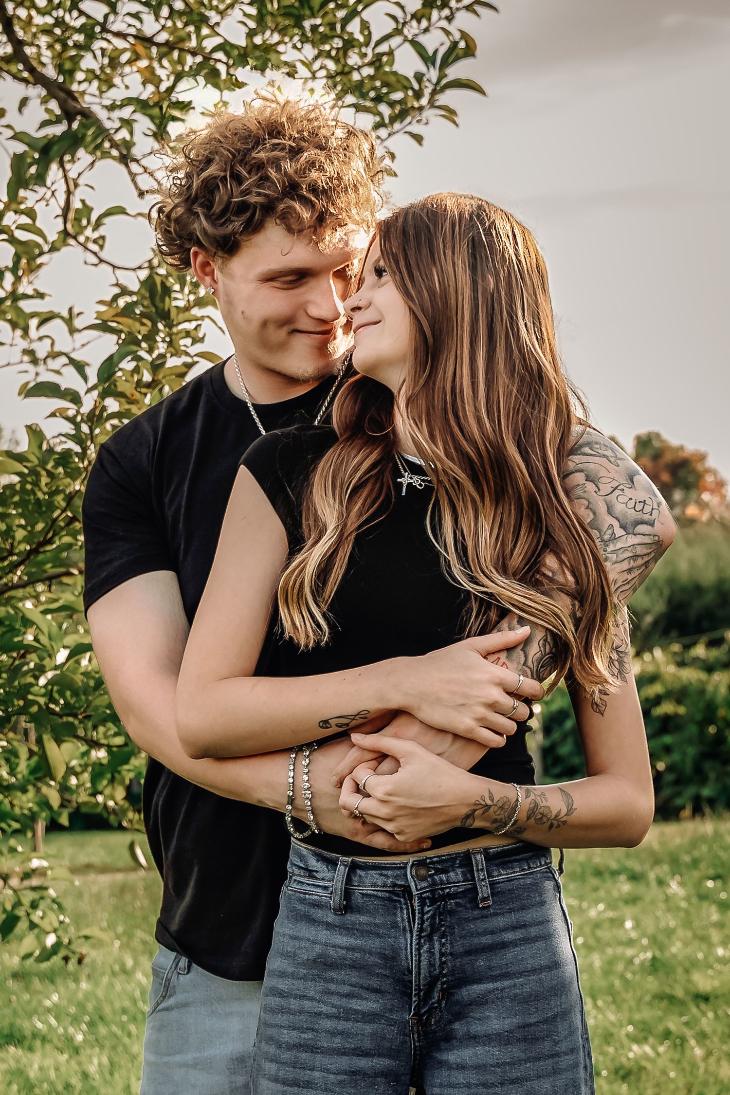 A young couple embracing and almost kissing outdoors, both wearing black tops, with green trees in the background.