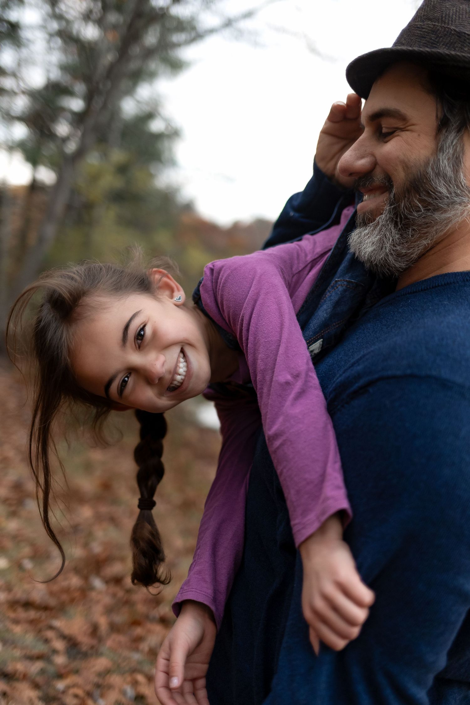 Laughing girl with braids being playfully carried by bearded father wearing hat in autumn forest.