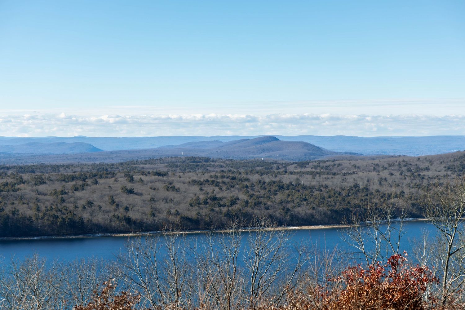 Scenic aerial view of blue lake surrounded by rolling hills and autumn trees under clear blue sky.