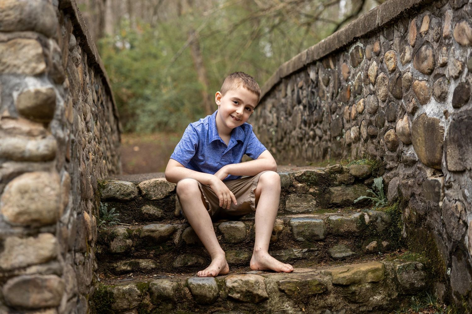 Smiling boy sitting barefoot on mossy stone steps outdoors, wearing blue shirt and tan shorts, surrounded by a stone wall.