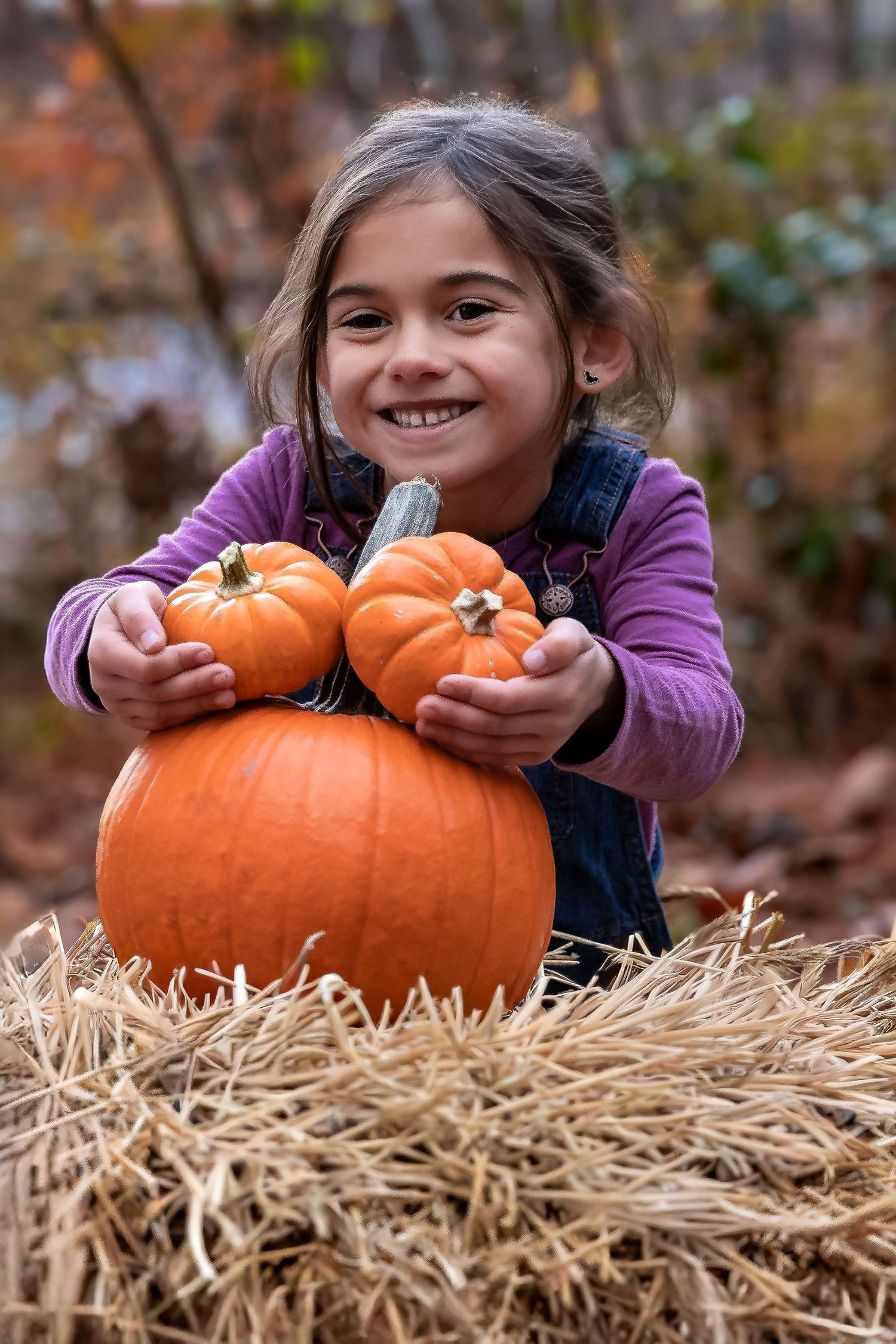 Smiling young girl hugging orange pumpkins on a hay bale in an autumn outdoor setting.