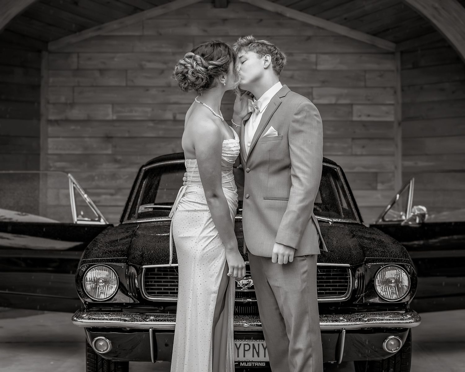 Couple sharing a romantic kiss in formal attire beside a classic Ford Mustang in black and white.
