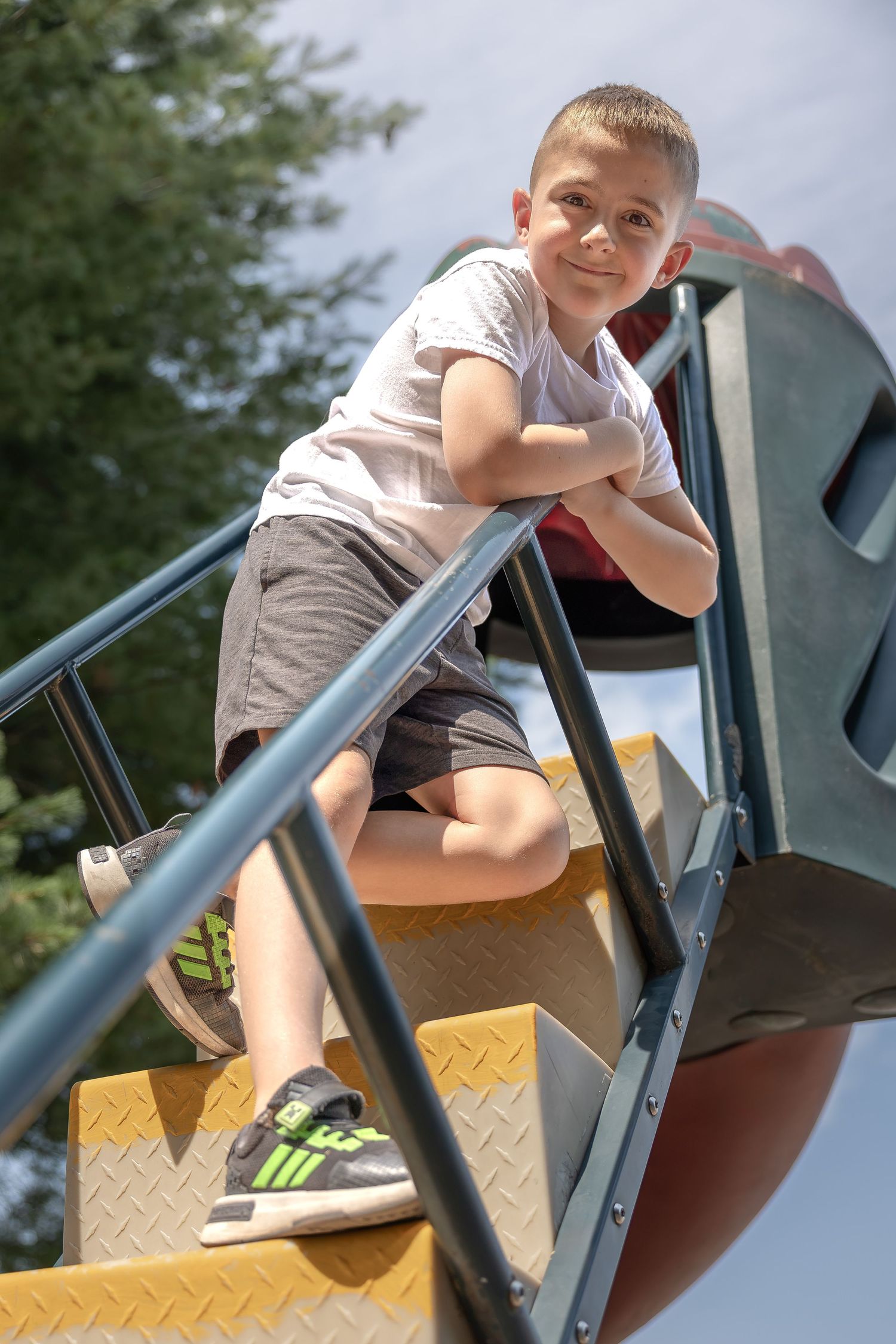 A young boy smiles while leaning on a railing at the top of playground stairs on a sunny day.