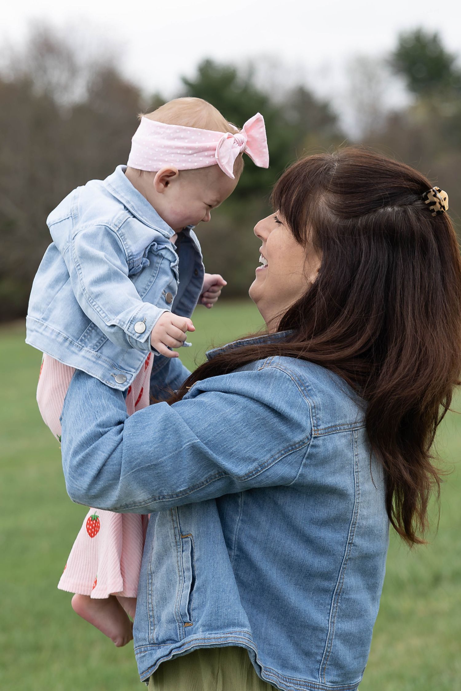 A smiling woman in a denim jacket holds up a baby girl wearing a pink bow headband outdoors.