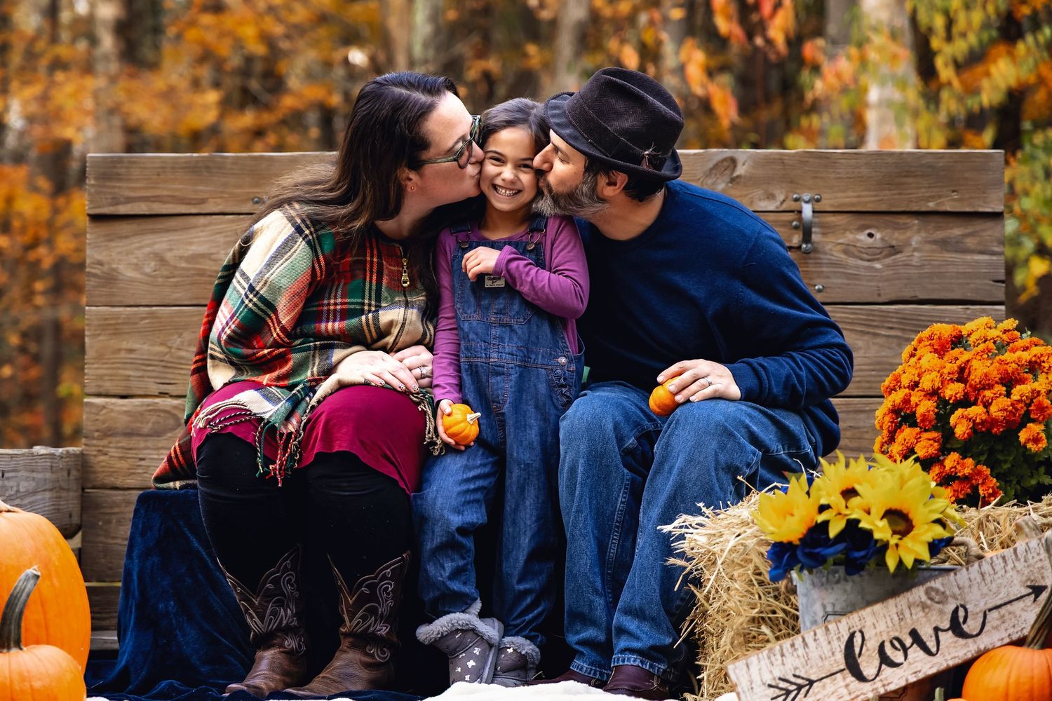 Family fall photo session with parents kissing child on cheeks, pumpkins and sunflowers in background.