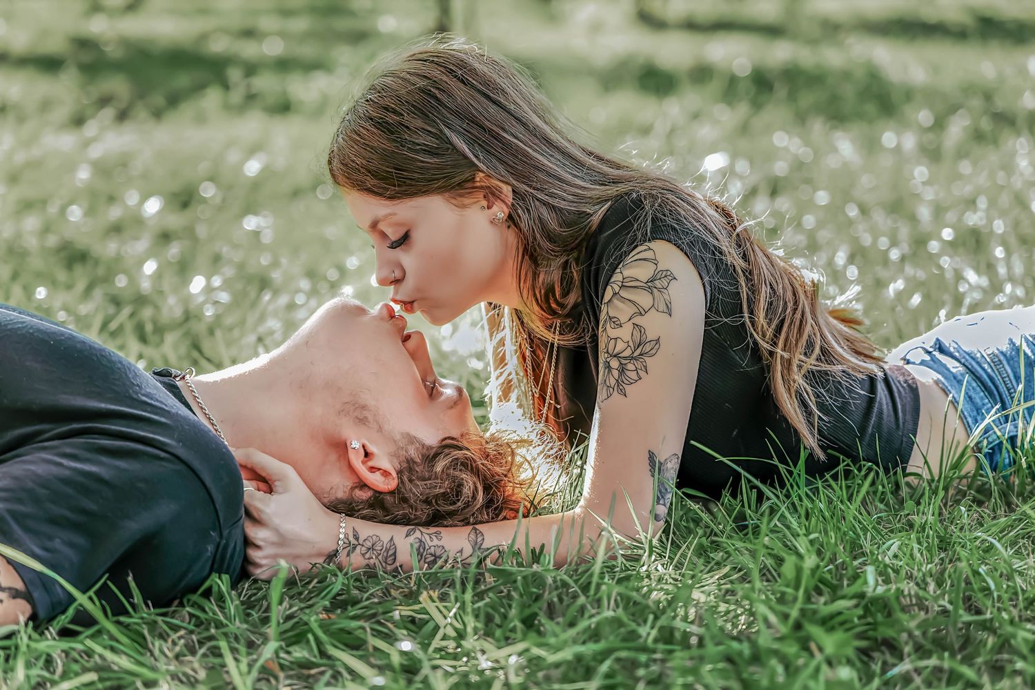 Couple with tattoos sharing a romantic kiss while lying in a sunny green grass field outdoors.