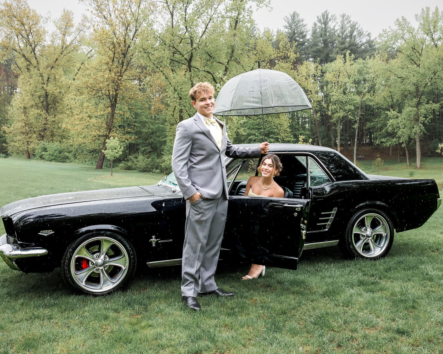 A couple poses beside a classic black Ford Mustang on a green lawn, man in gray suit holding an umbrella.