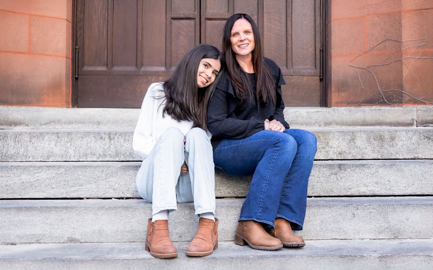 Two women smiling and sitting together on stone steps in front of a wooden door, wearing casual jeans and boots.