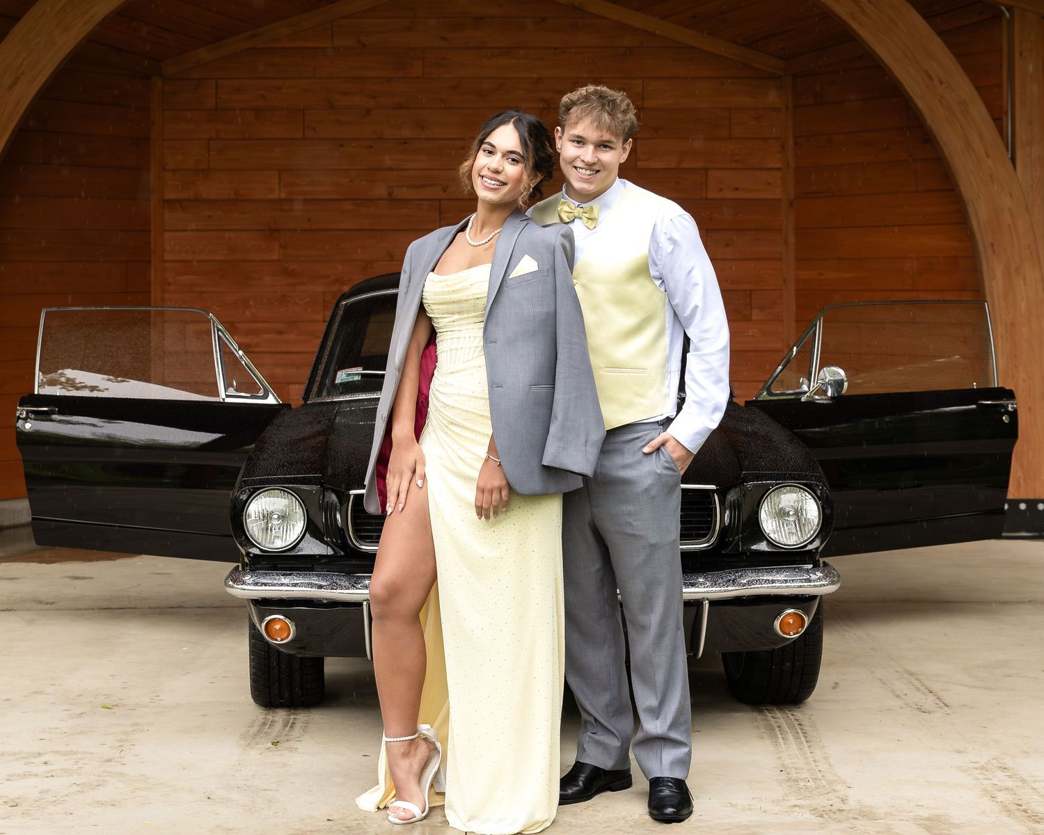 Prom couple posing in formal attire beside a classic black Ford Mustang in front of a wooden building.