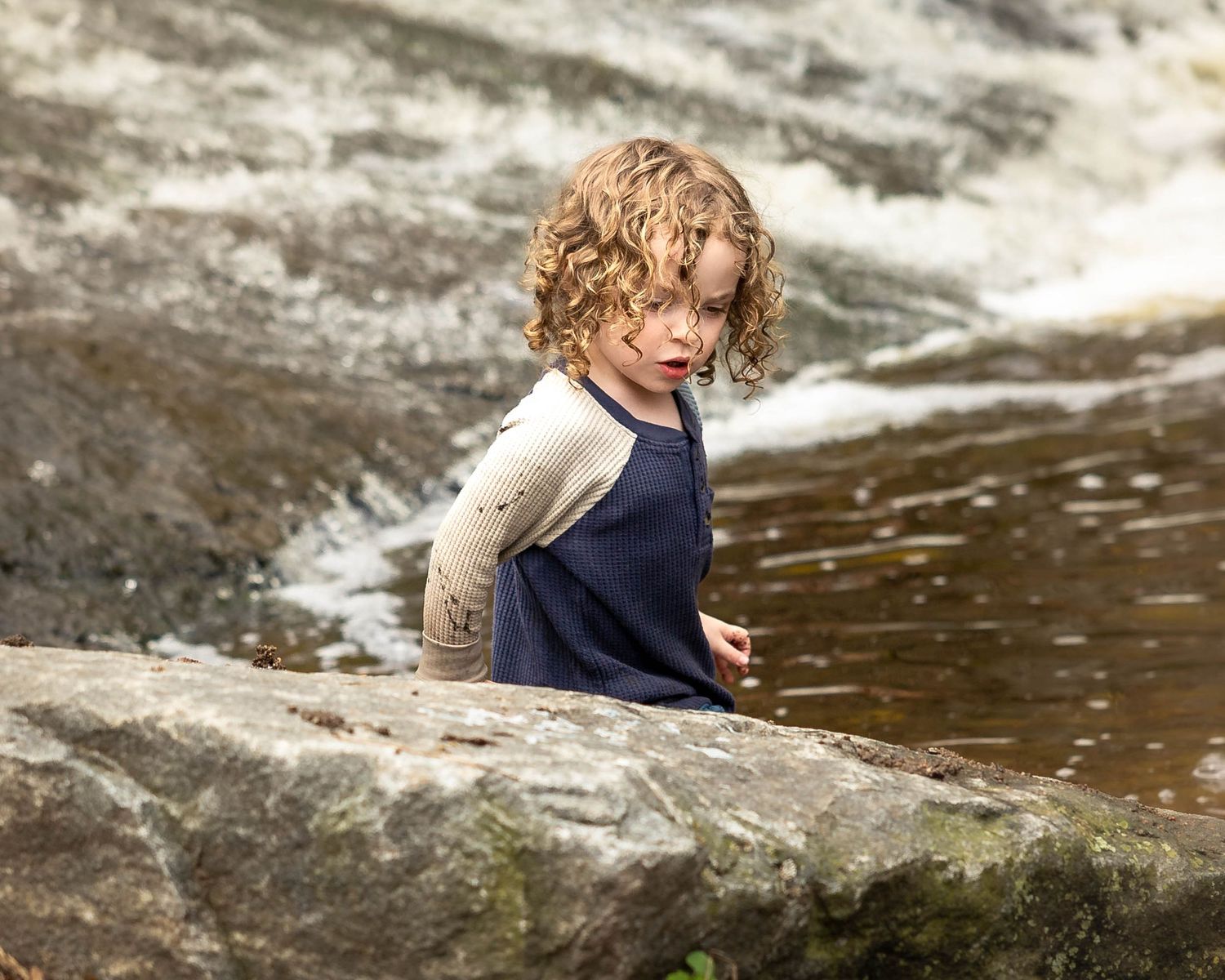Curly-haired child in navy swimsuit exploring rocky riverbank with rushing water in background.