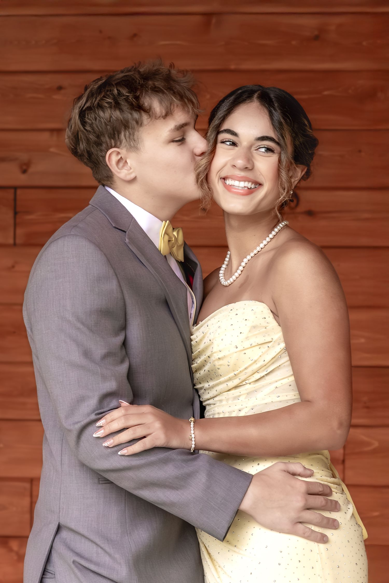 A young couple in formal prom attire, boy in gray suit kisses girl in yellow strapless gown wearing pearl jewelry.