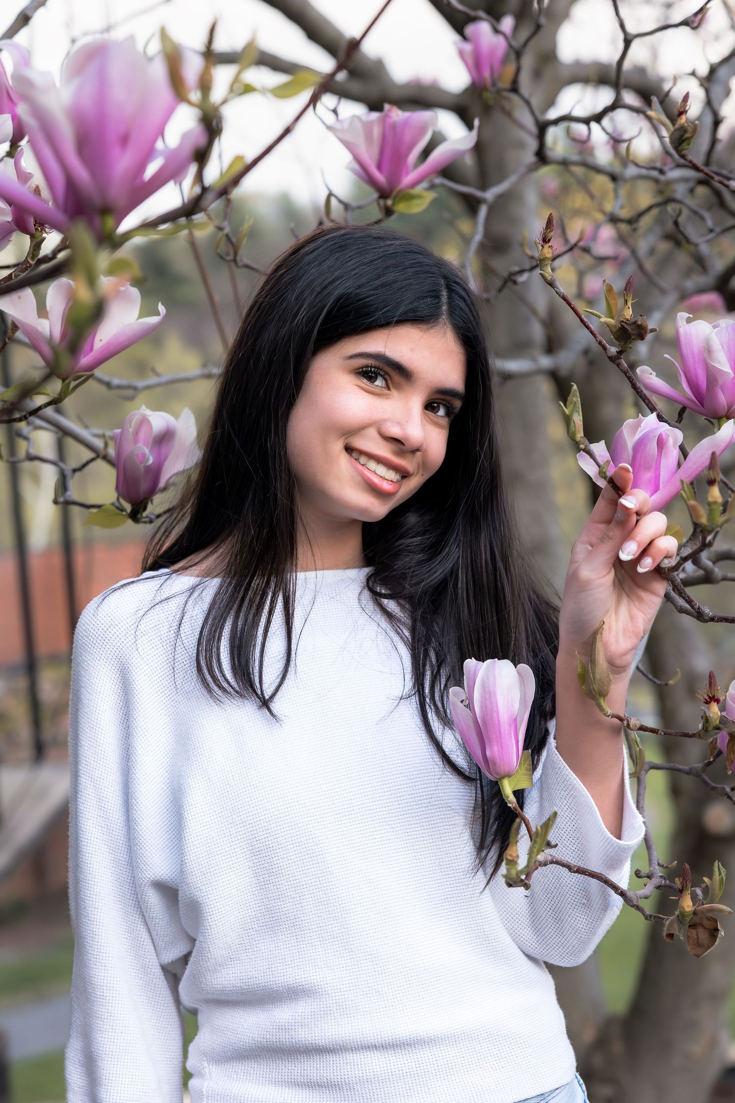 Young woman with dark hair smiling among pink magnolia blossoms, wearing a white sweater in spring garden.