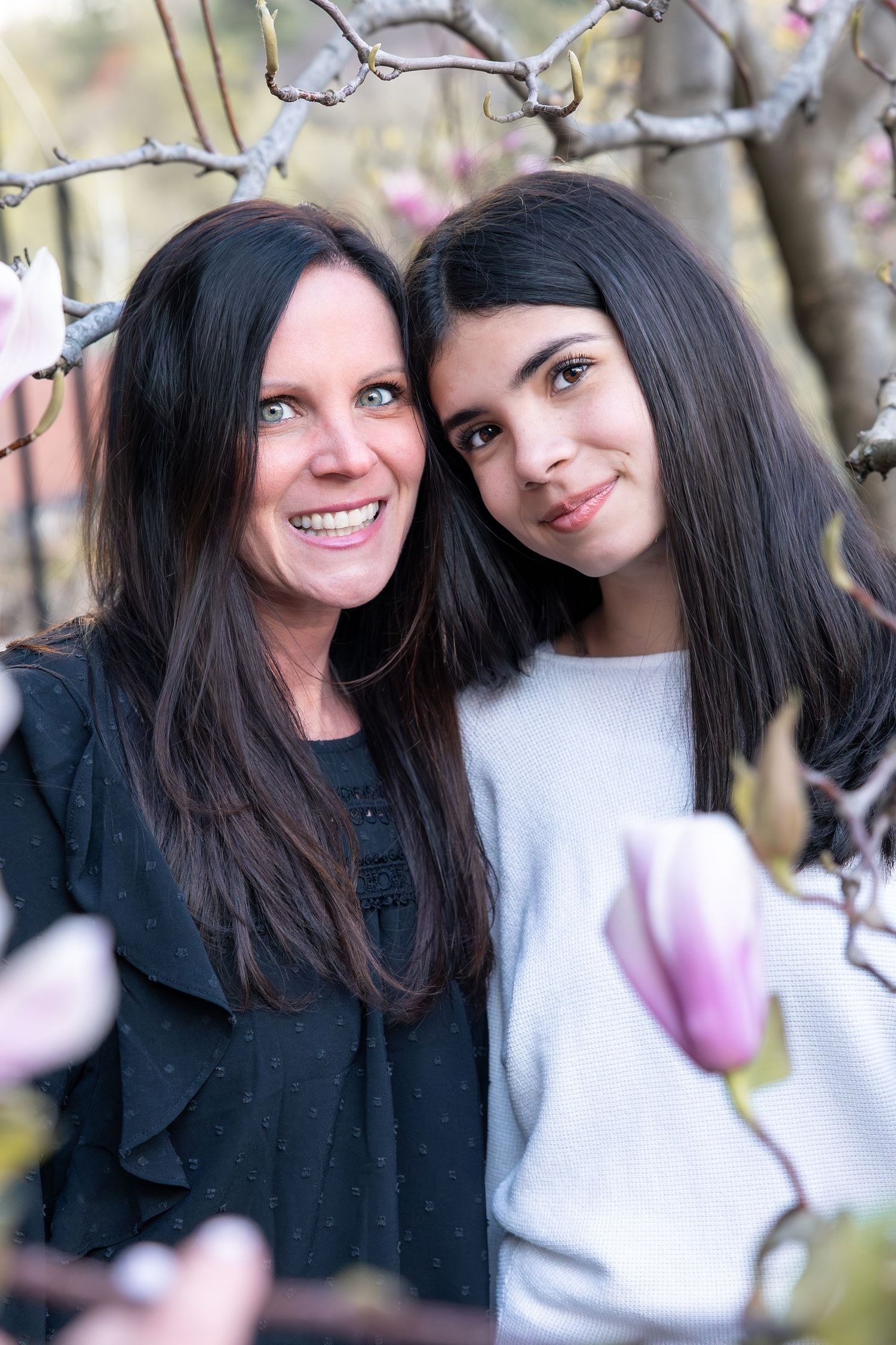 A smiling mother and daughter pose together outdoors near blooming magnolia tree branches in spring.
