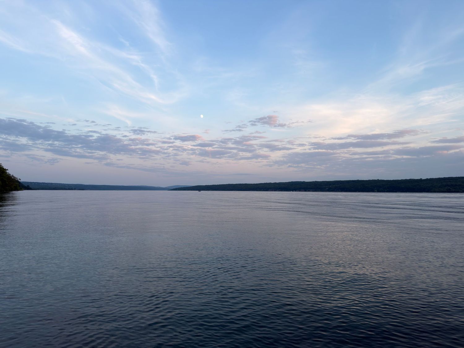 Calm lake at dusk with soft reflections, wispy clouds, and forested shoreline under a pastel blue sky.