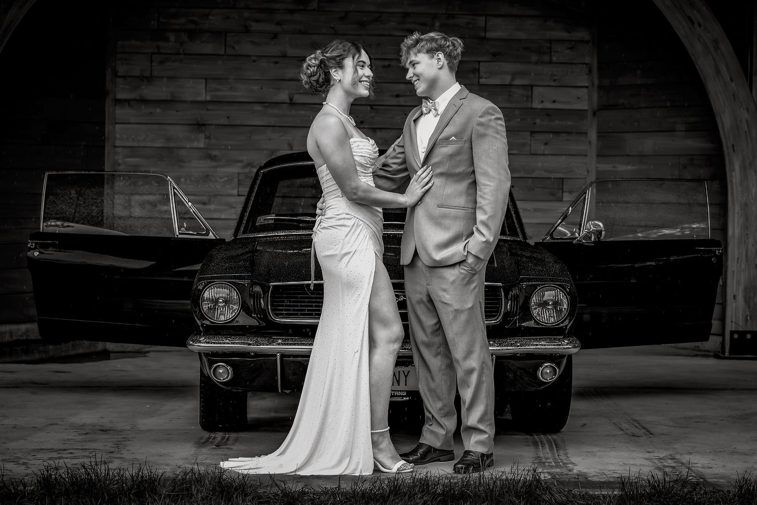 Black and white wedding photo of bride and groom embracing in front of a classic car.