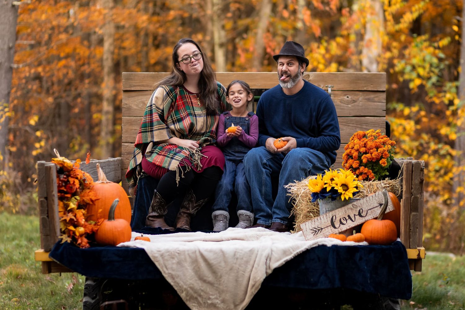 Family of three smiling on rustic truck bed with fall pumpkins, mums, and colorful autumn foliage backdrop.