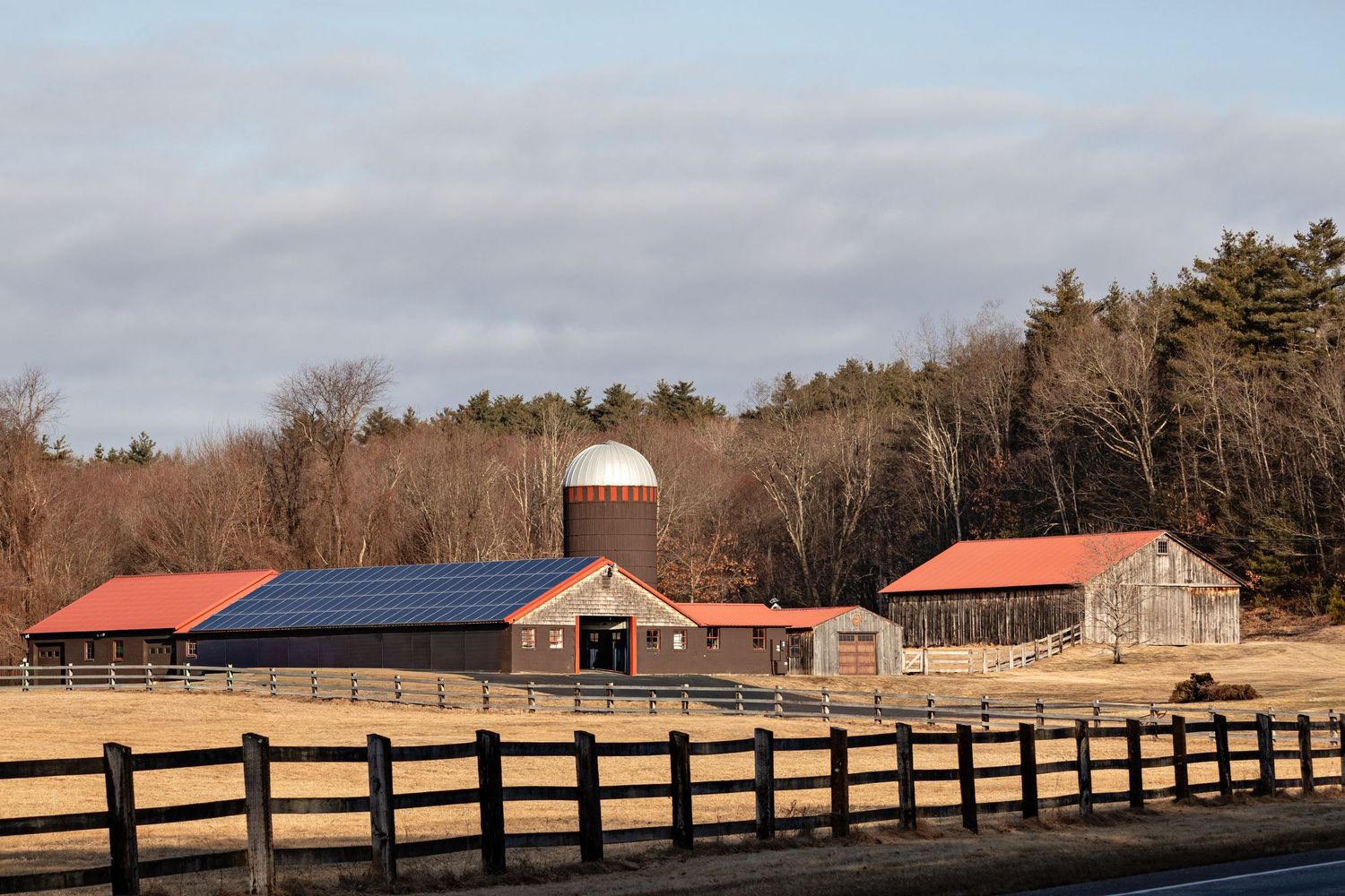 Rustic farm with red-roofed barn, solar panels, silo, and wooden fence surrounded by bare trees under cloudy skies.