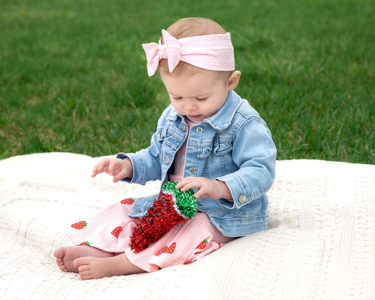 Baby girl in denim jacket and pink strawberry dress wearing bow headband plays with toy on white blanket outdoors.