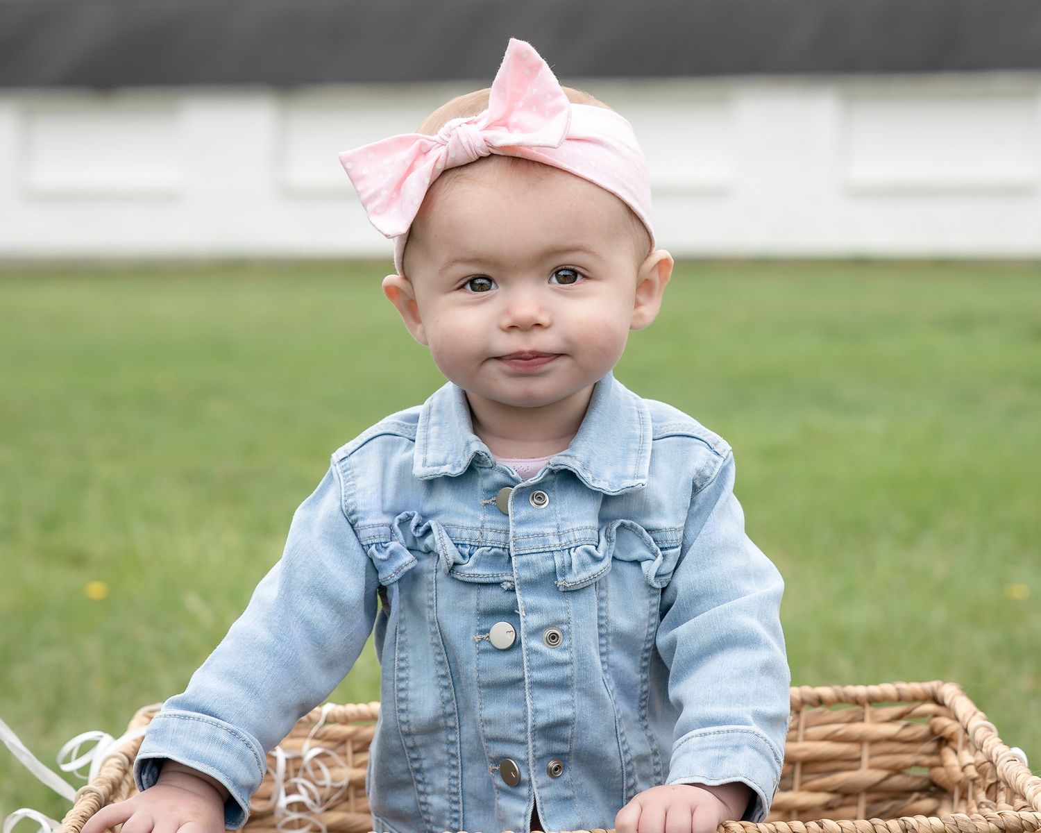 Smiling baby girl in denim jacket and pink bow headband sitting in wicker basket outdoors on green grass.