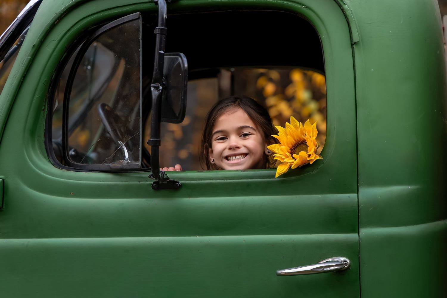 Smiling girl holding sunflowers peers through the window of a vintage green truck in an autumn setting.