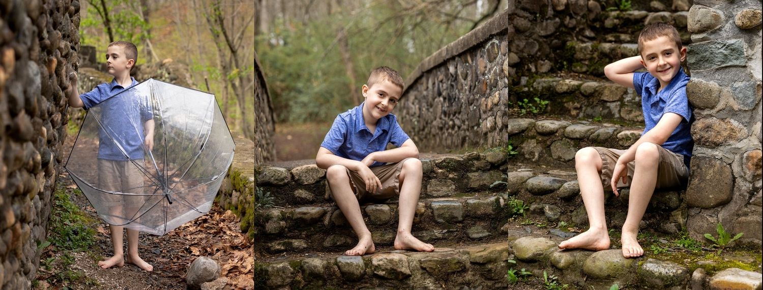 A young boy poses barefoot outdoors near stone steps and trees, holding a clear umbrella in a blue shirt.