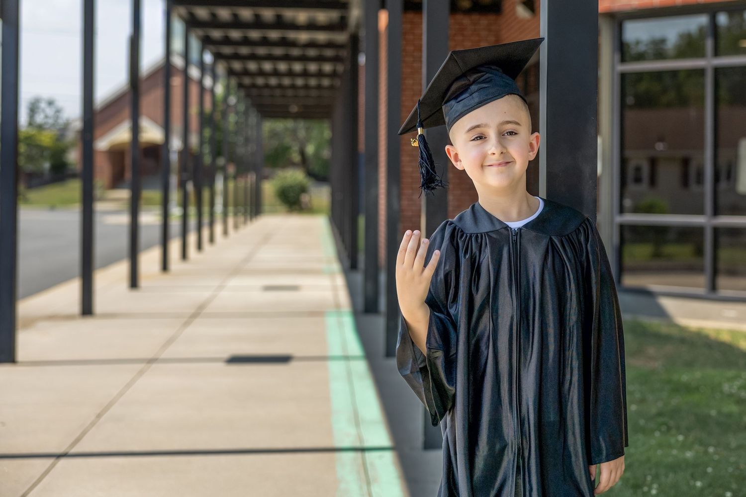Young child in black graduation cap and gown smiling outdoors near school building on a sunny day.