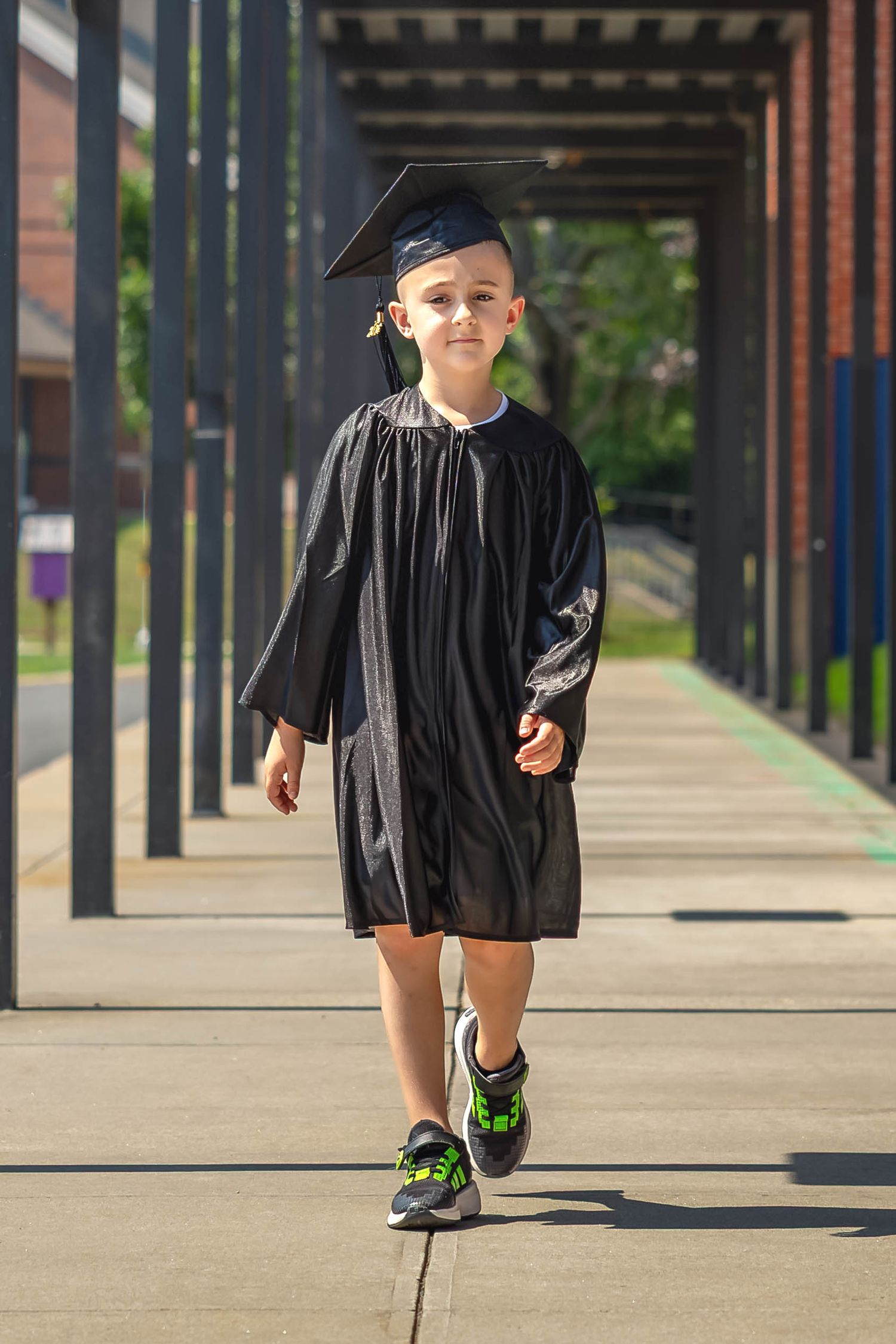 Young boy wearing black graduation cap and gown walks confidently down a covered school walkway in sneakers ready to attend a new school