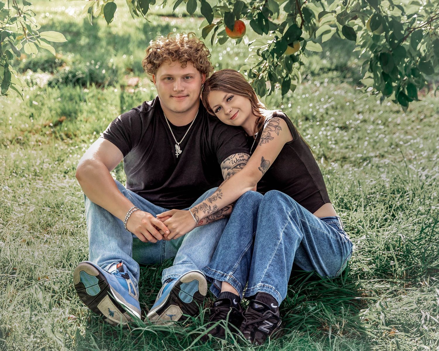 Young couple in matching black shirts and jeans sitting together on grass under a fruit tree outdoors.