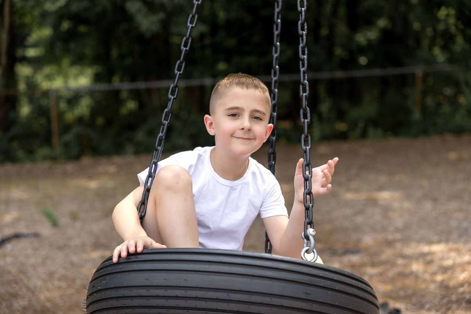 Young boy smiling on a tire swing at a playground, wearing a white t-shirt, surrounded by trees.