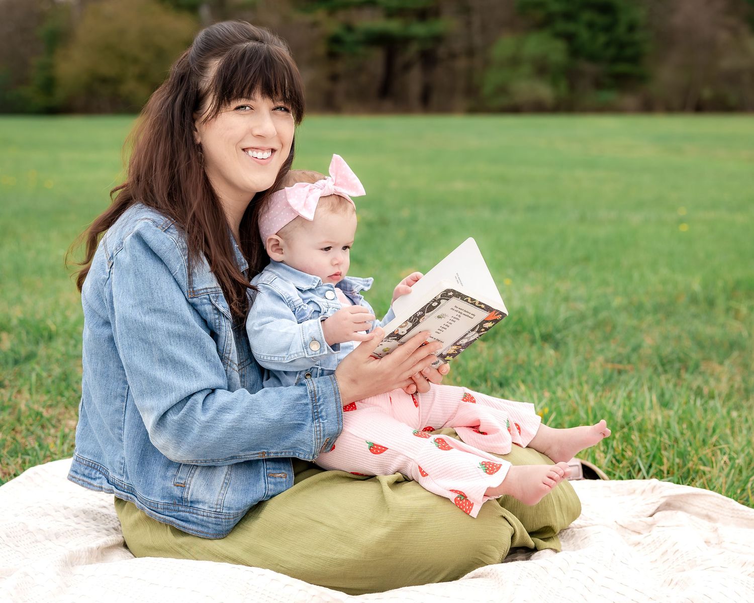 Mother sitting on blanket in park reading book with baby girl in pink bow and floral outfit.