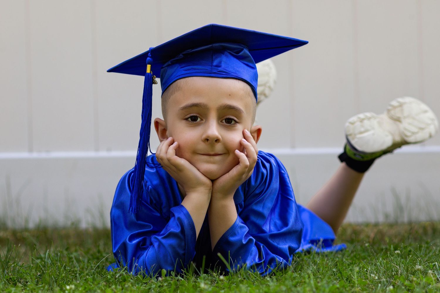 Young boy in blue graduation cap and gown lying on grass, smiling with hands on cheeks.