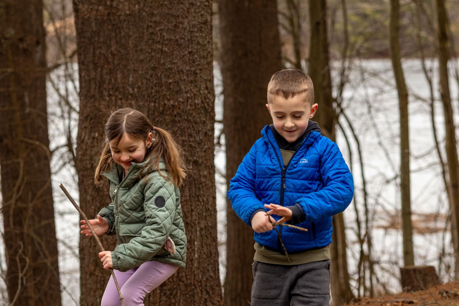 Two children playing in a snowy forest, girl in green jacket holding stick, boy in blue puffer jacket exploring outdoors.