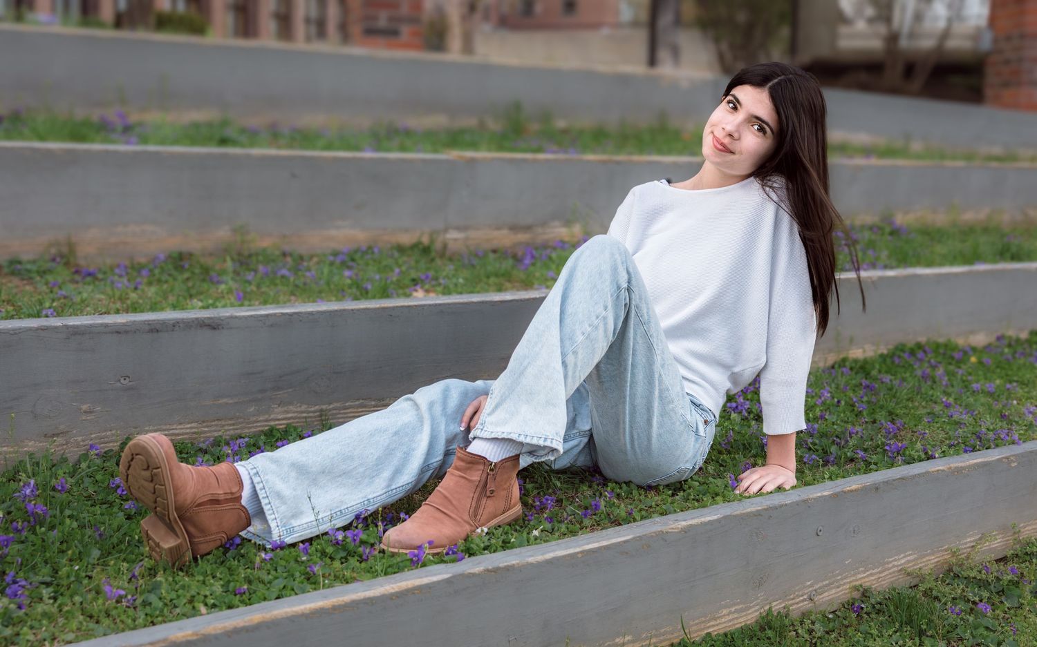 Young woman in white sweater, jeans, and brown boots sitting on outdoor steps with purple flowers.