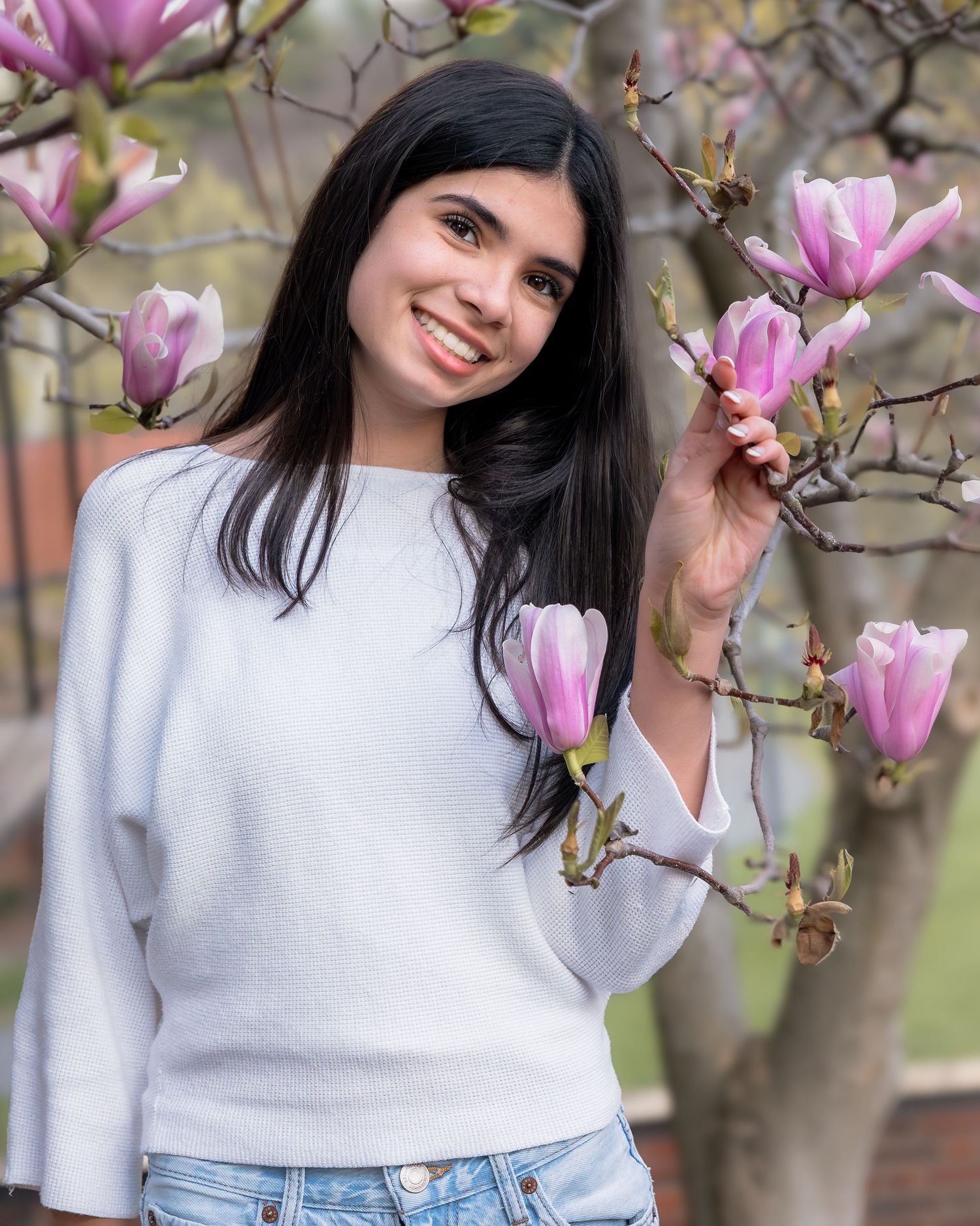 Young woman in white sweater smiling among pink magnolia blossoms in spring garden.