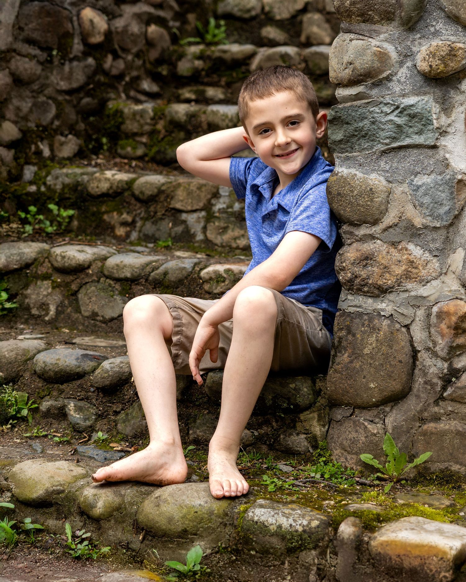 A smiling boy in a blue shirt and khaki shorts sits barefoot on stone steps against a rustic rock wall outdoors.