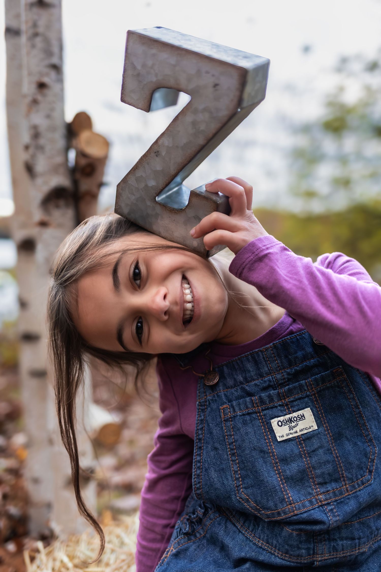 A cheerful young girl in denim overalls holds a metal letter Z while posing outdoors in an autumn setting.