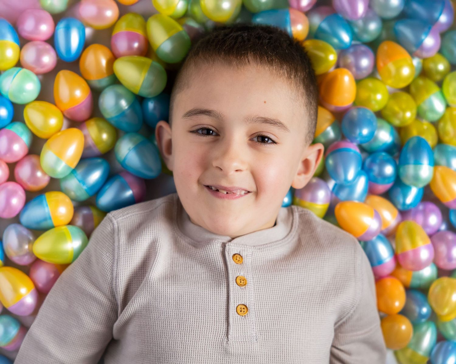 Smiling young boy lying among colorful Easter eggs in pink, blue, yellow, and green, wearing a gray henley shirt.
