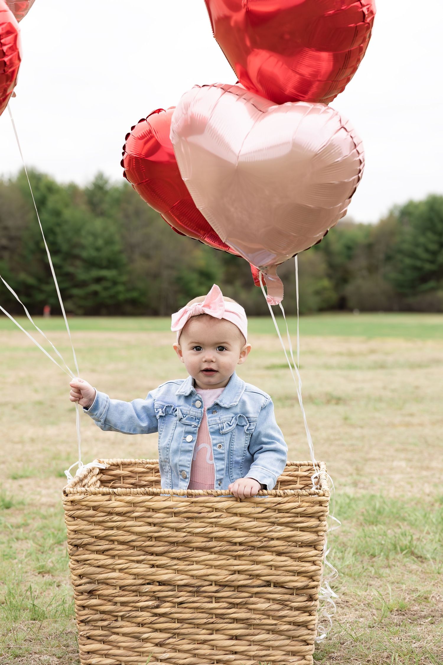 Baby girl in denim jacket stands in wicker basket with red and pink heart-shaped balloons outdoors.