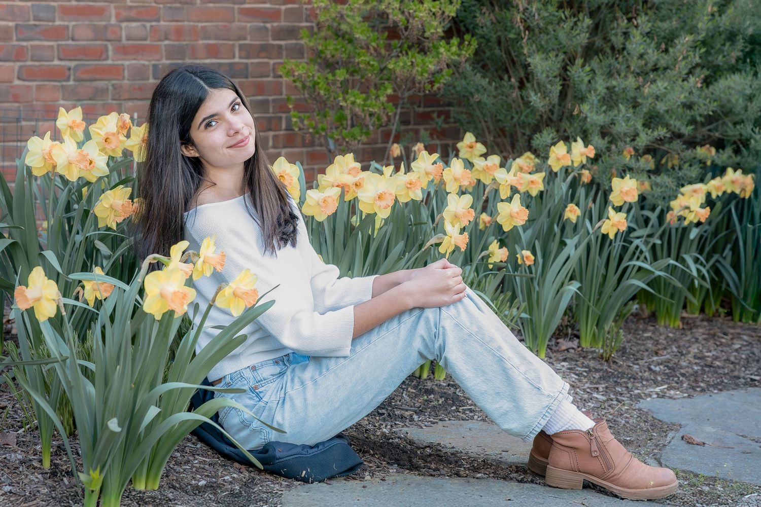 Young woman sitting among yellow daffodils by a brick wall, wearing white top and light jeans with tan boots.