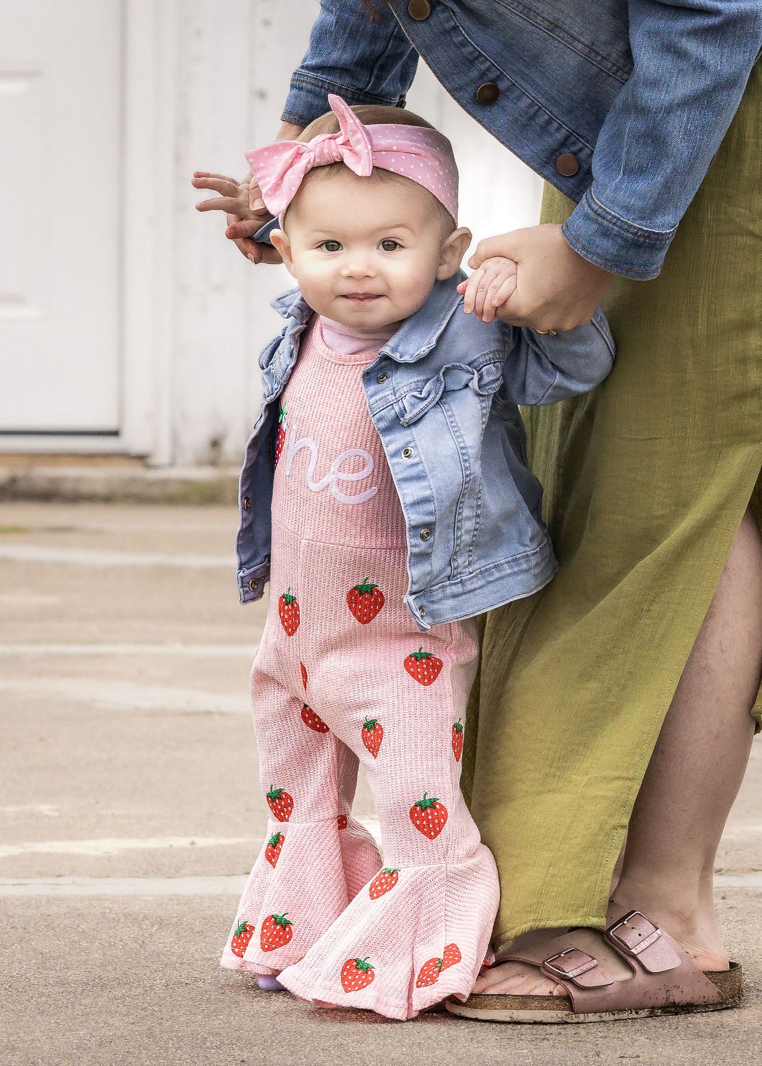 Adorable baby girl in strawberry bell-bottom pants, denim jacket, and pink bow headband, standing with adult support.