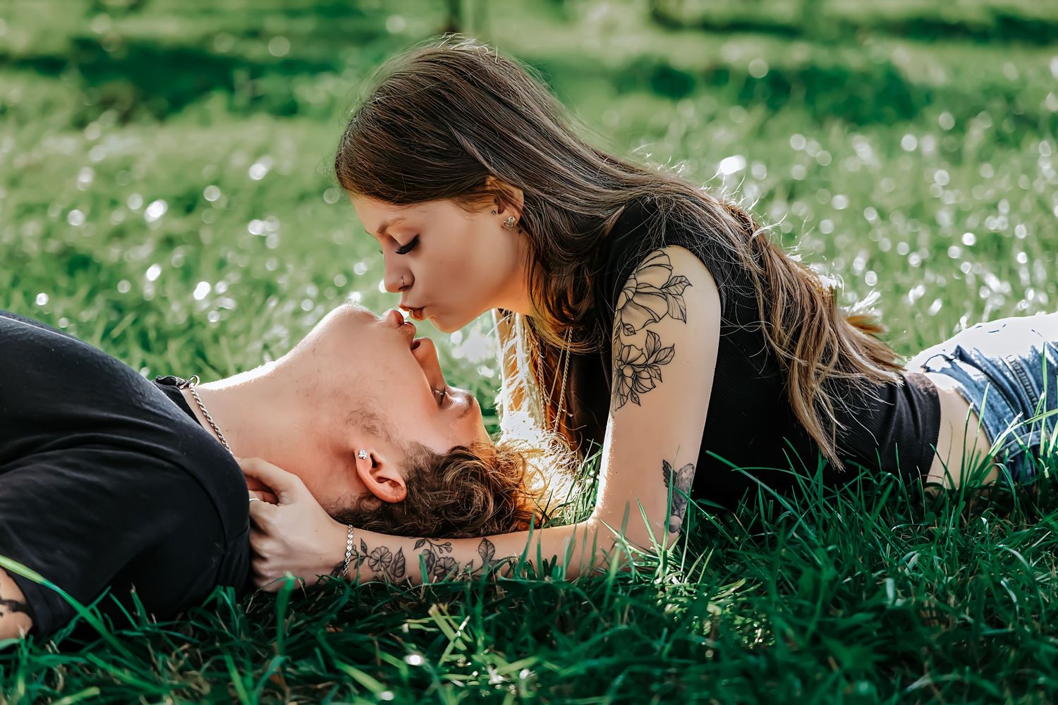 A couple sharing a romantic kiss while lying in lush green grass outdoors, both with visible tattoos.