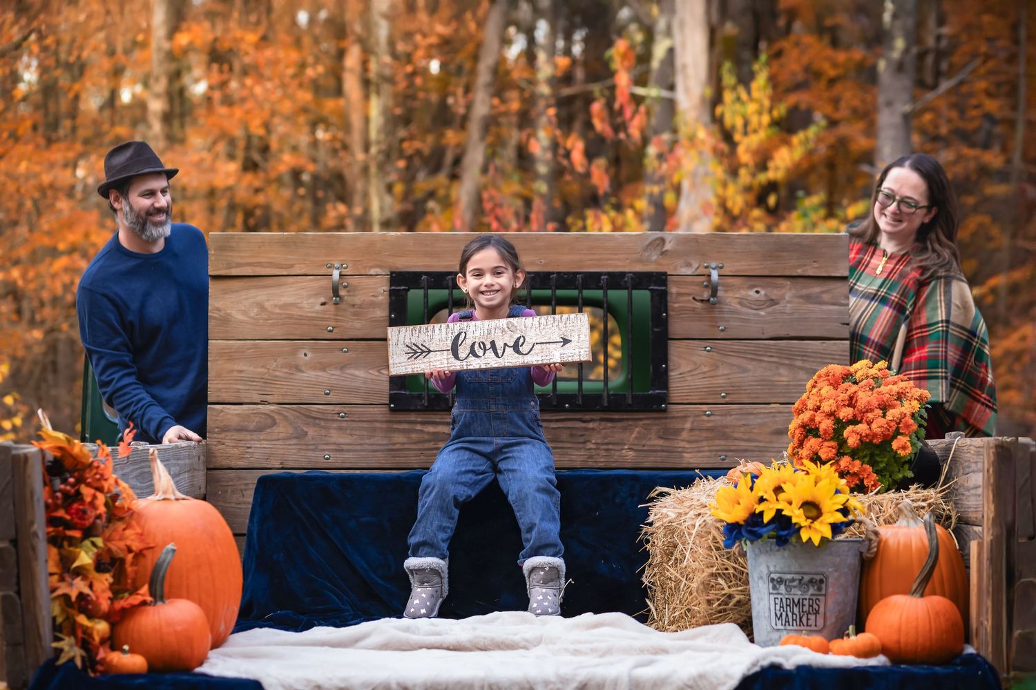 Family of three posing at a rustic fall harvest display with pumpkins, hay bales, and autumn foliage backdrop.