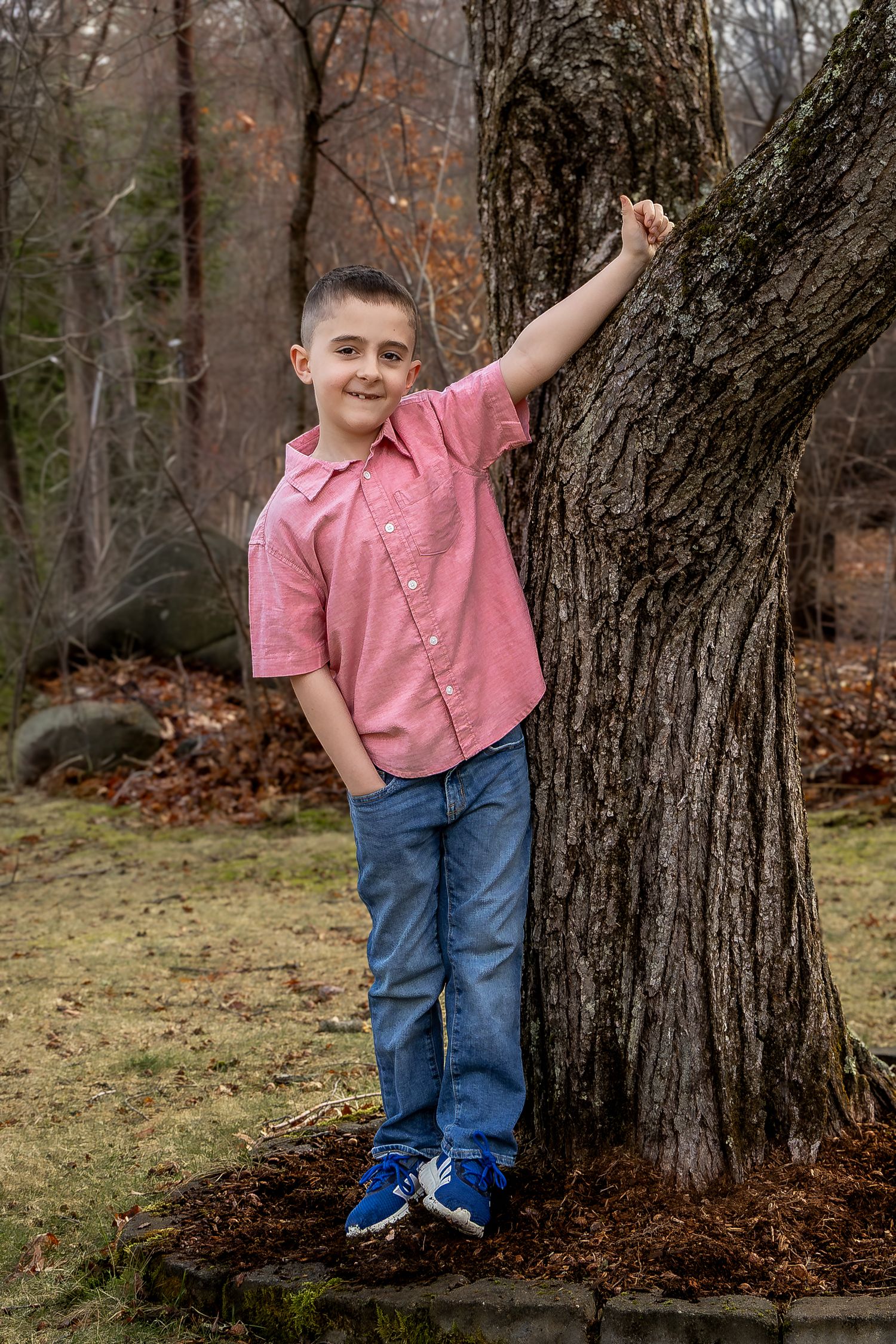 A young boy in a pink shirt and jeans leans against a large tree in an outdoor wooded setting.