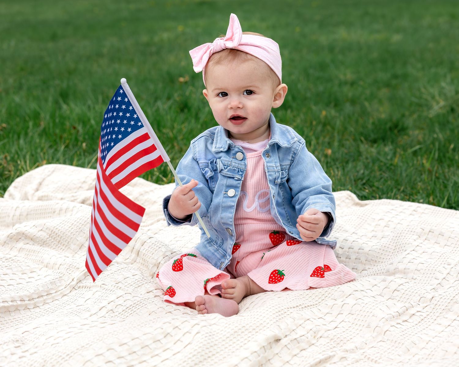 Baby girl in denim jacket and strawberry leggings holds American flag while sitting on white blanket in grass.